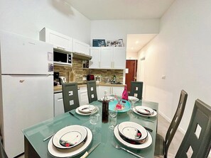 Een eethoek met een glazen tafel, gedekt voor vier personen, omringd door stoelen. Links is een witte koelkast en een magnetron, en boven de aanrecht een houten kast.