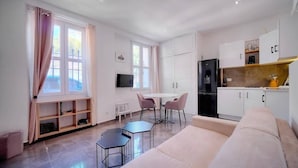 Ein modernes Wohnzimmer mit Sofa, Fernseher, Esstisch mit Stühlen und einer Küchenzeile mit Schränken und Kühlschrank.