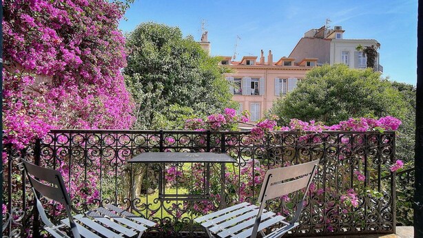 Een balkon met een ijzeren hekwerk, twee metalen stoelen, een kleine tafel en uitzicht op een gebouw met roze bloemen en groen.