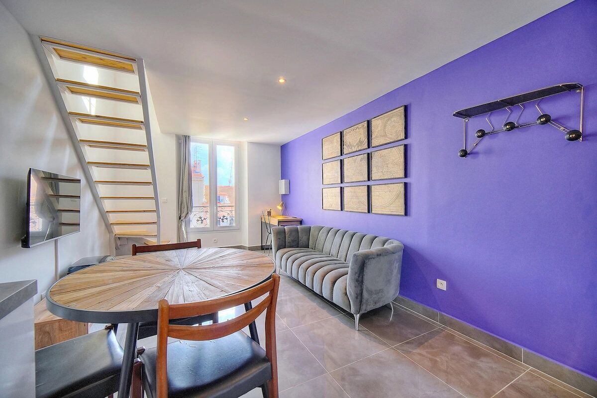 A dining area with a round wooden table and chairs, a grey sofa, a purple wall with framed artwork, and a metal ladder.