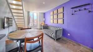 Ein Essbereich mit einem runden Holztisch und Stühlen, einem grauen Sofa, einer violetten Wand mit gerahmten Kunstwerken und einer Metallleiter.
