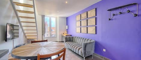 A dining area with a round wooden table and chairs, a grey sofa, a purple wall with framed artwork, and a metal ladder.