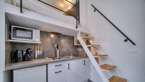 Une cuisine compacte équipée d’un four à micro-ondes, d’une machine à café et d’ustensiles de cuisine posés sur le plan de travail, d’un petit réfrigérateur et d’un escalier blanc à marches en bois.