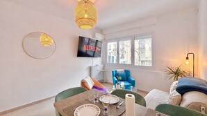 Een moderne woonkamer met een eettafel, een blauwe fauteuil, een bank en een televisie die aan de muur is bevestigd, plus een groot raam.