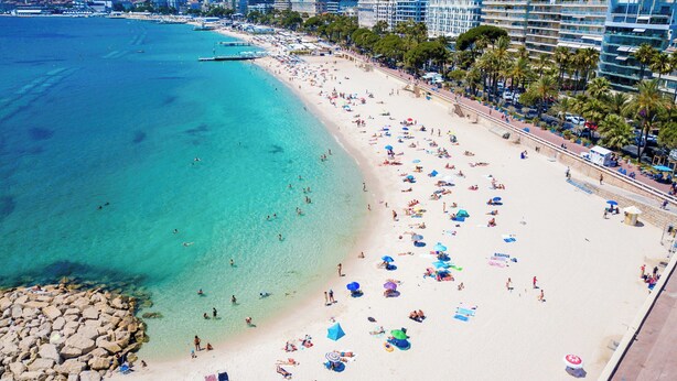 Een strand met helder turquoise water, een rotsachtige kustlijn en een promenade met gebouwen en palmbomen.