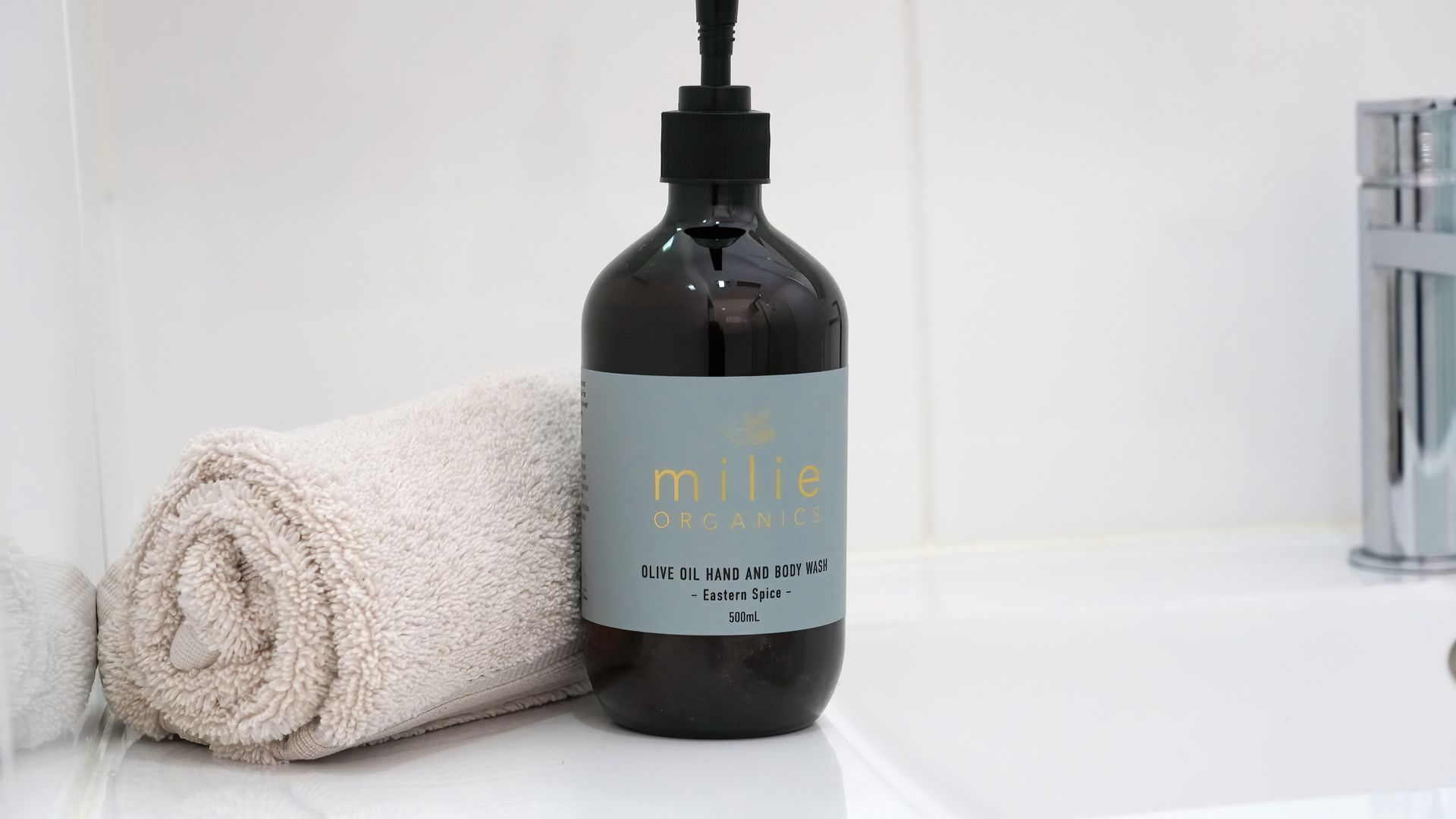 A bottle of organic olive oil hand and body wash with a pump dispenser, placed next to a rolled towel on a white surface.