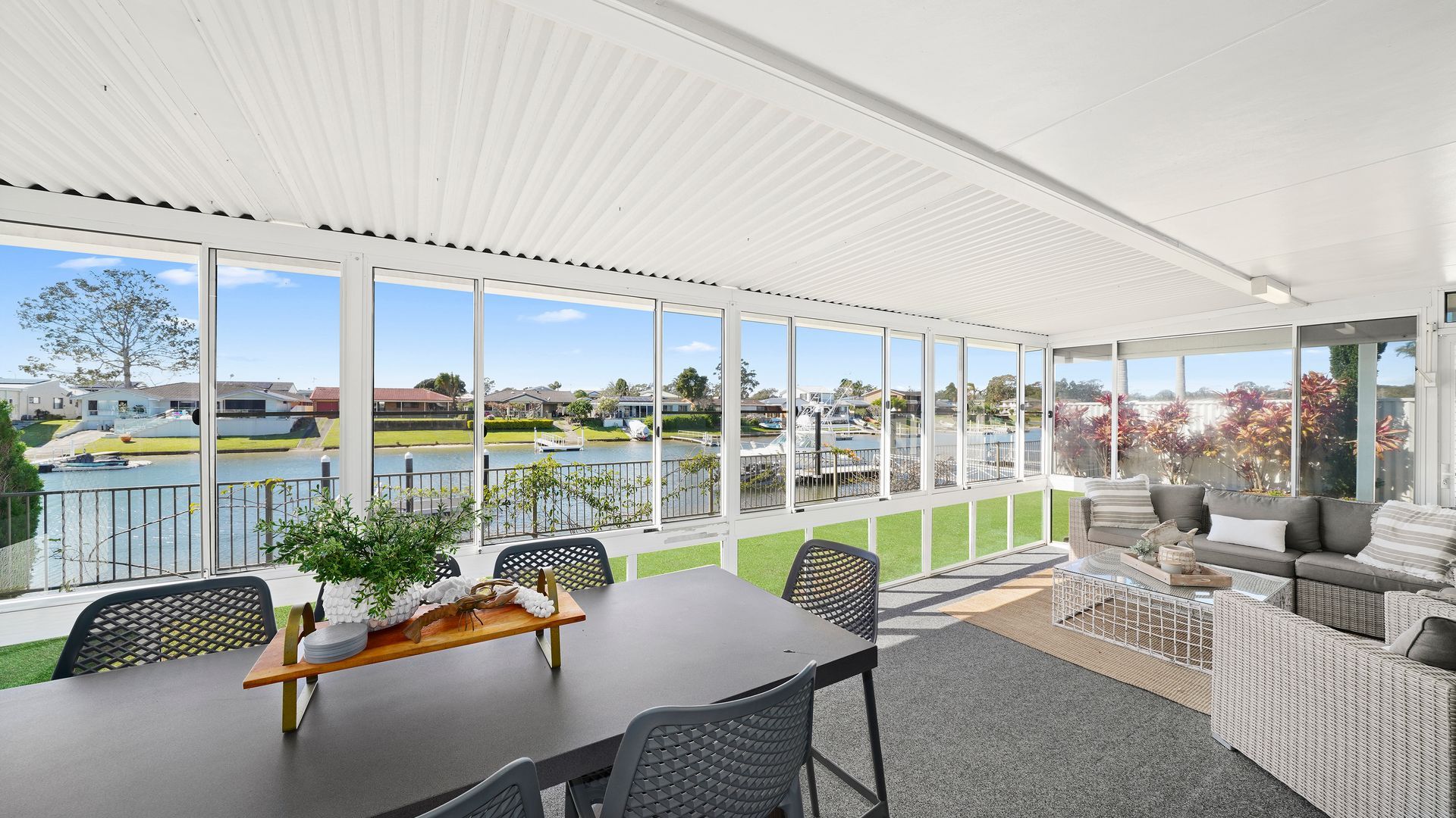 A covered outdoor dining area with a view of a lake and boats, furnished with a table, chairs, and a sofa.