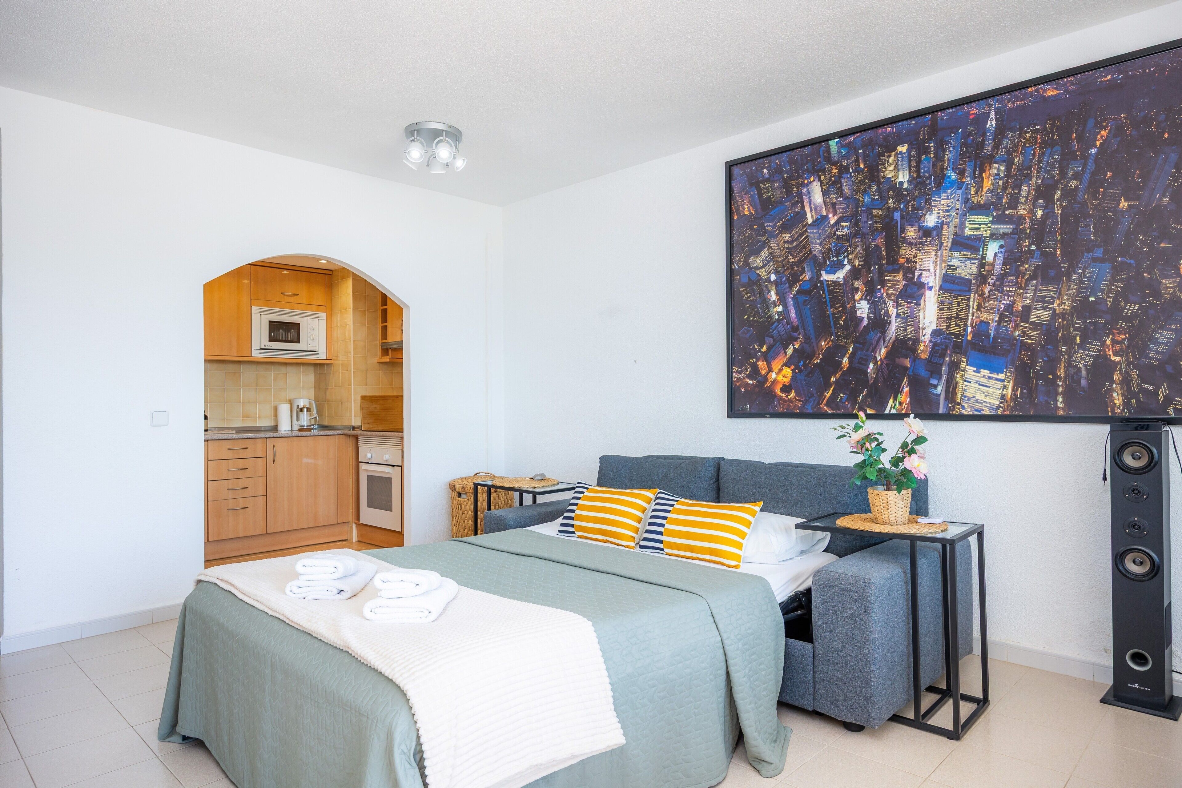 Un dormitorio moderno con cama, una mesita de noche pequeña, un cuadro grande con un paisaje urbano y una kitchenette al fondo.