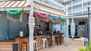 Un bar frente a la playa con mostrador de piedra, taburetes y un cartel que prohíbe botellas de vidrio.