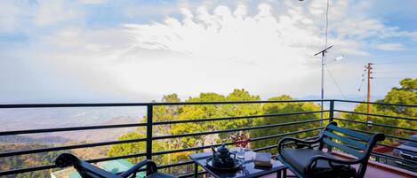 Un balcón con dos bancos, una mesa con tetera y libro, y vistas a un paisaje verde.