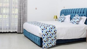 Un dormitorio con cabecera tapizada en azul, cama con ropa de cama blanca y un plaid estampado, dos lámparas de noche y una mesita de noche con un teléfono.