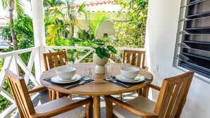 Een ronde houten tafel met vier stoelen, een potplant en witte kommen en glazen op een balkon met een witte reling en tropische planten op de achtergrond.