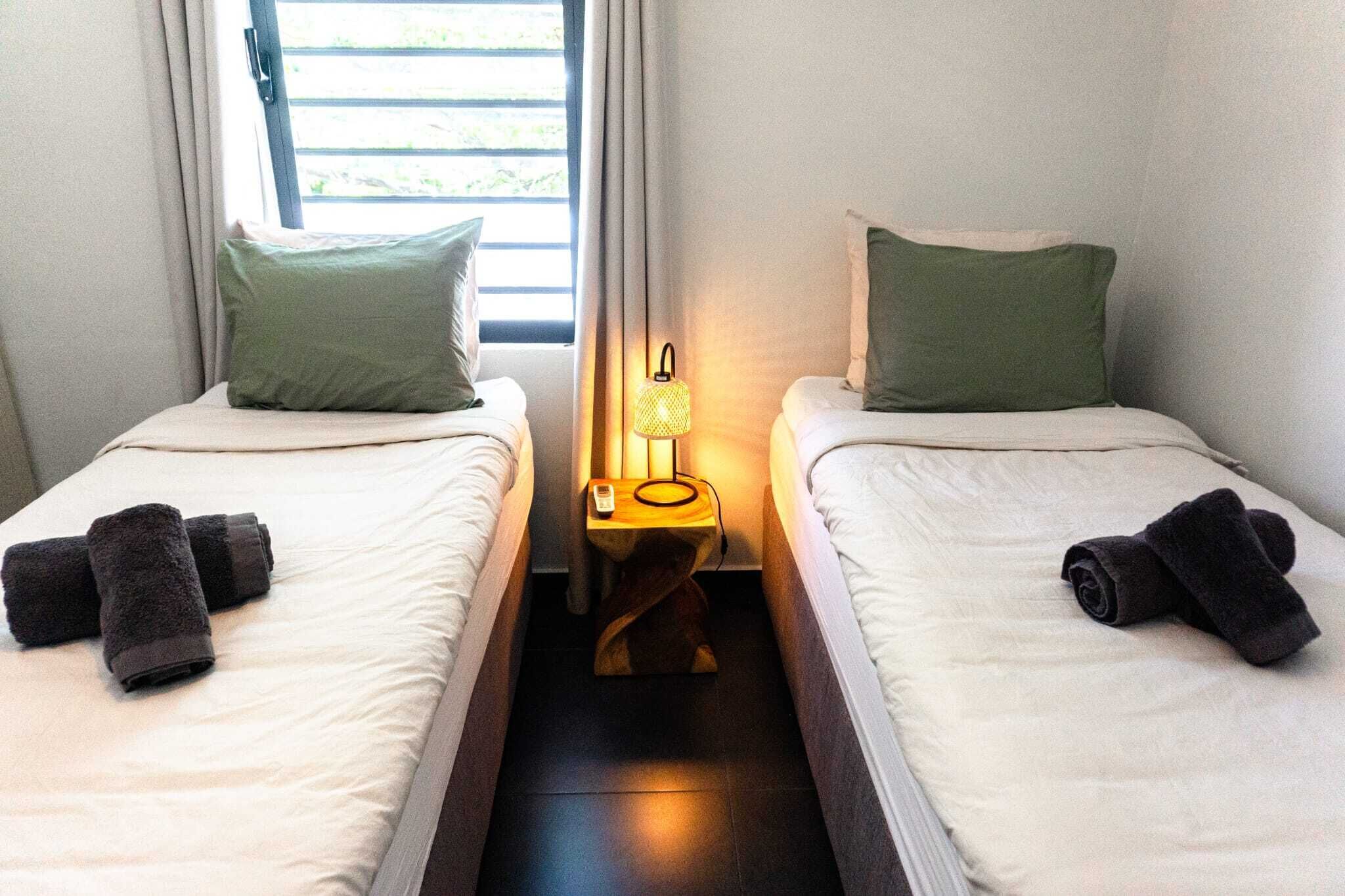 Twee enkele bedden met groene kussens, een nachtkastje met een lamp, en een raam met gordijnen.
