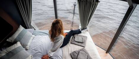 Una cama con vista al mar, techo de cristal y una ventana con cortinas.