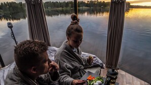 Ett par sitter vid ett bord och äter, med utsikt över en sjö och en skog, och på bordet står mat och dryck.