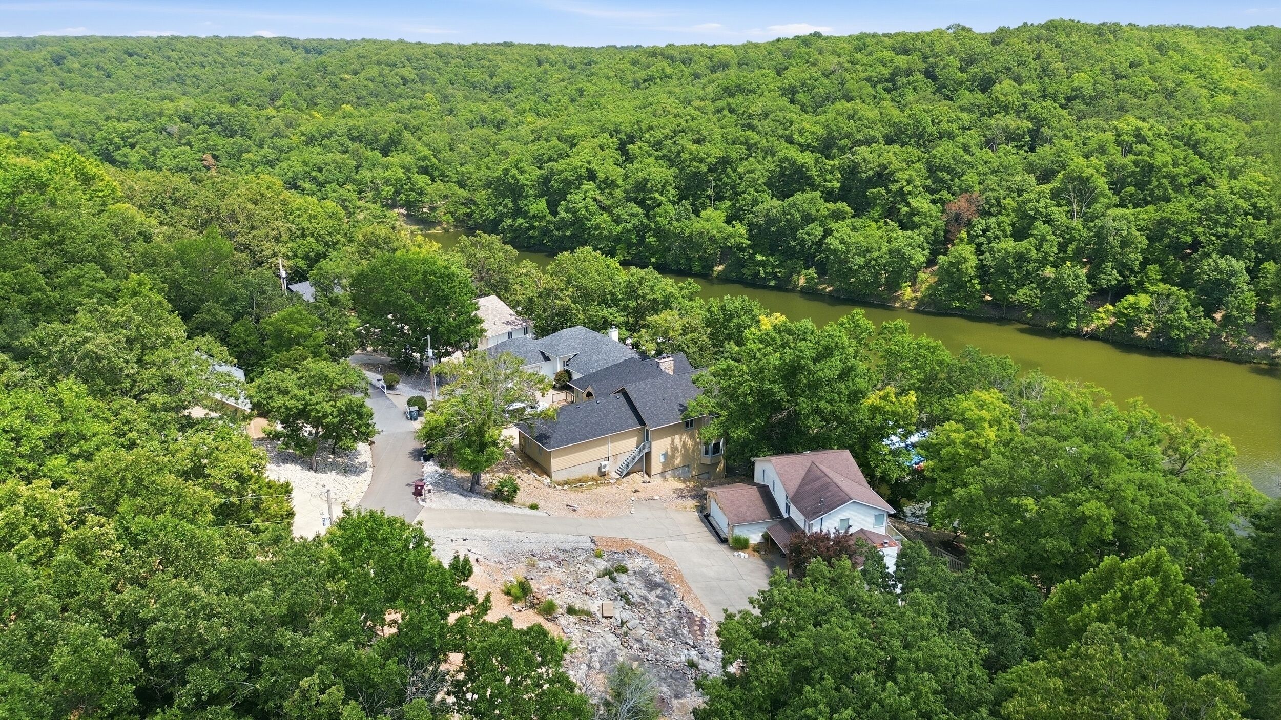 A residential area with a house surrounded by dense forest, a clear sky, and a river.
