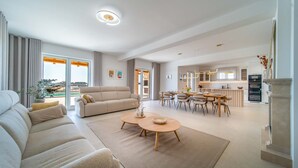 Ein modernes Wohnzimmer mit einer Couch, einem Couchtisch und einem Essbereich. Im Hintergrund befindet sich eine Küche mit Esstisch und Stühlen.