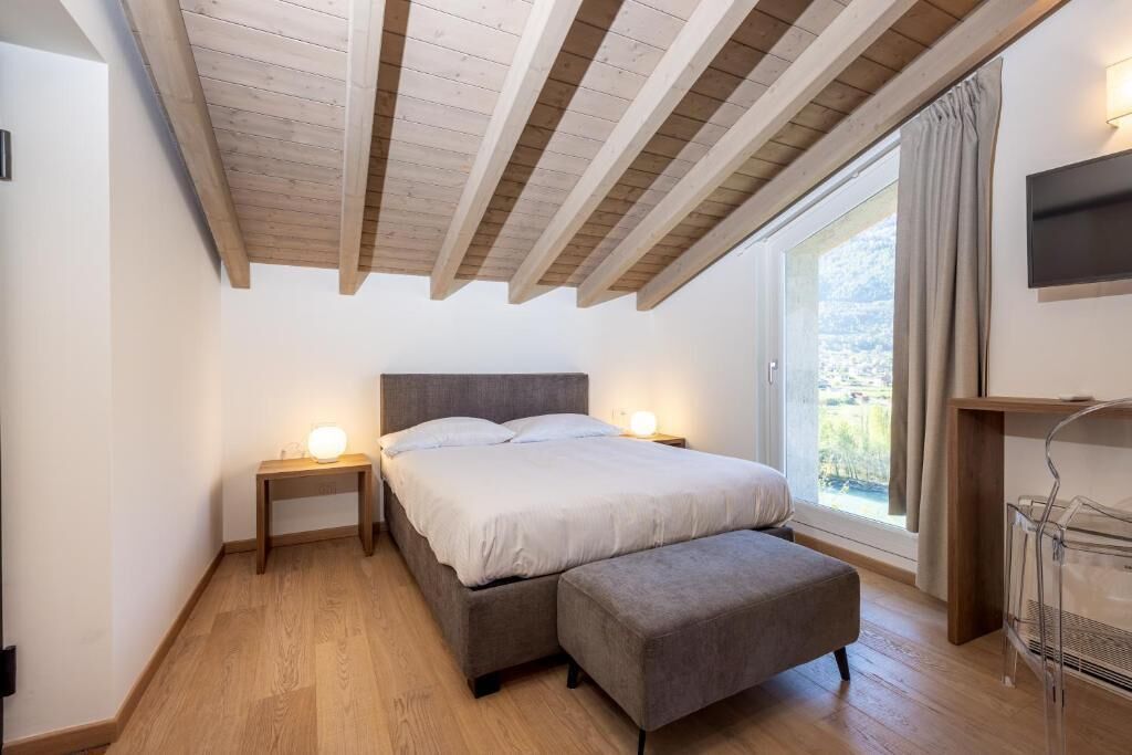 Un dormitorio con cama, mesitas de noche, un banco, televisor y ventana con cortinas.