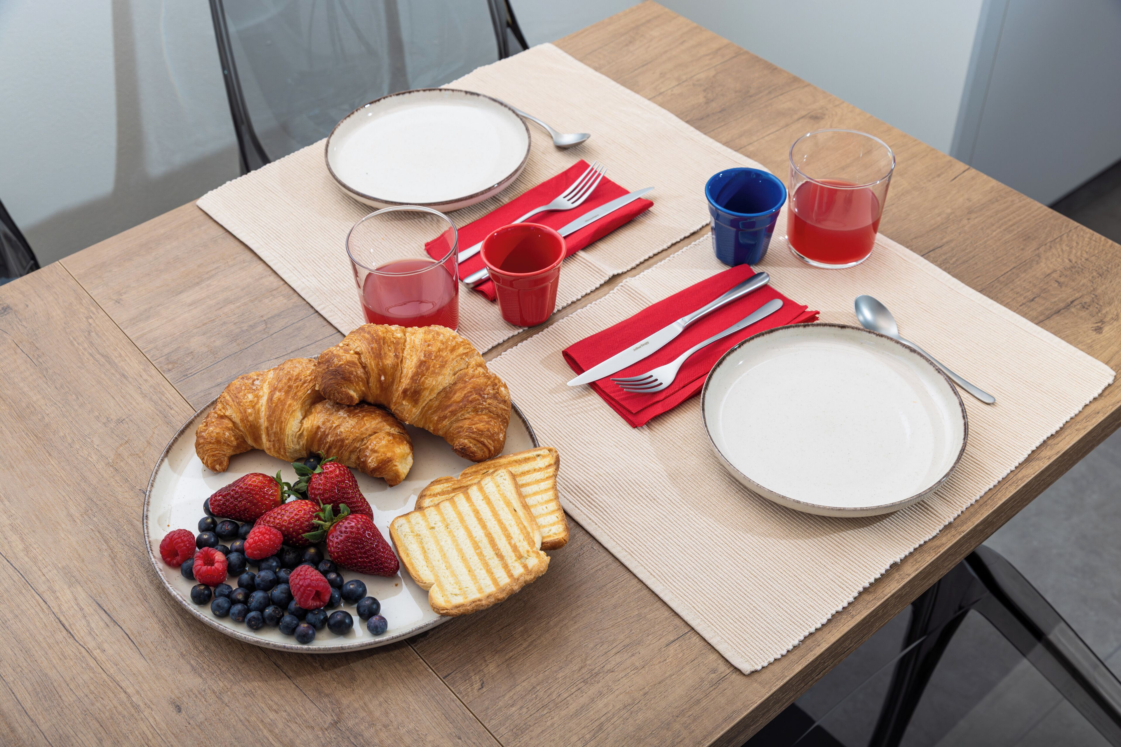 Une table en bois dressée pour le petit-déjeuner, avec une assiette contenant des croissants, des fraises et des myrtilles, un verre de jus rouge et une tasse de thé.