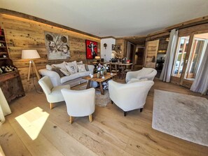 Ein gemütliches Wohnzimmer mit einer Holzwand, einer Couch, zwei Sesseln, einem Couchtisch und einem Essbereich im Hintergrund.