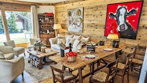 Een gezellige woonkamer met een houten eettafel, stoelen en een bank. Een boekenplank met diverse spullen en een schilderij van een koe aan de muur.
