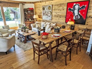 Ein gemütliches Wohnzimmer mit einem Holzesstisch, Stühlen und einer Couch. Ein Bücherregal mit diversen Gegenständen und ein Kuhbild an der Wand sind zu sehen.