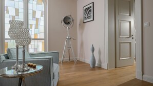 Une pièce comprenant un canapé, une table avec une lampe, un trépied, un tableau encadré et une porte.