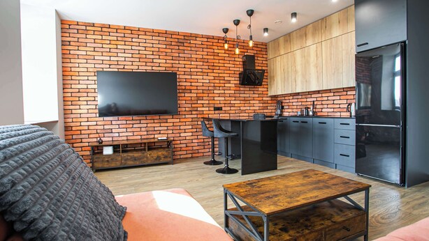 Un salon moderne avec un mur en briques, un téléviseur à écran plat, un canapé, une table basse et un coin cuisine équipé de placards et d’un réfrigérateur.