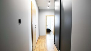 Un couloir étroit avec un plancher en bois, des murs blancs et une porte donnant sur une chambre à coucher où se trouve un lit.