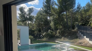 A modern outdoor pool area with a glass table and chairs, surrounded by a stone wall and trees. - Dominant house overlooking pine forest (Les Matelles)