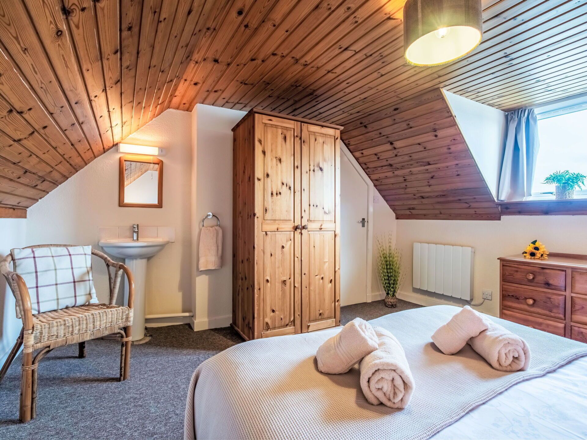 Quarto aconchegante com teto de madeira, cama com toalhas dobradas, guarda-roupa de madeira, cadeira de vime, pia e janela com cortinas.