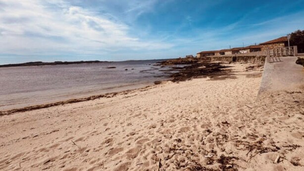 A beach with a sandy shore, a clear sky, and a building with a balcony on the right. - As Casoeiras (O Grove)
