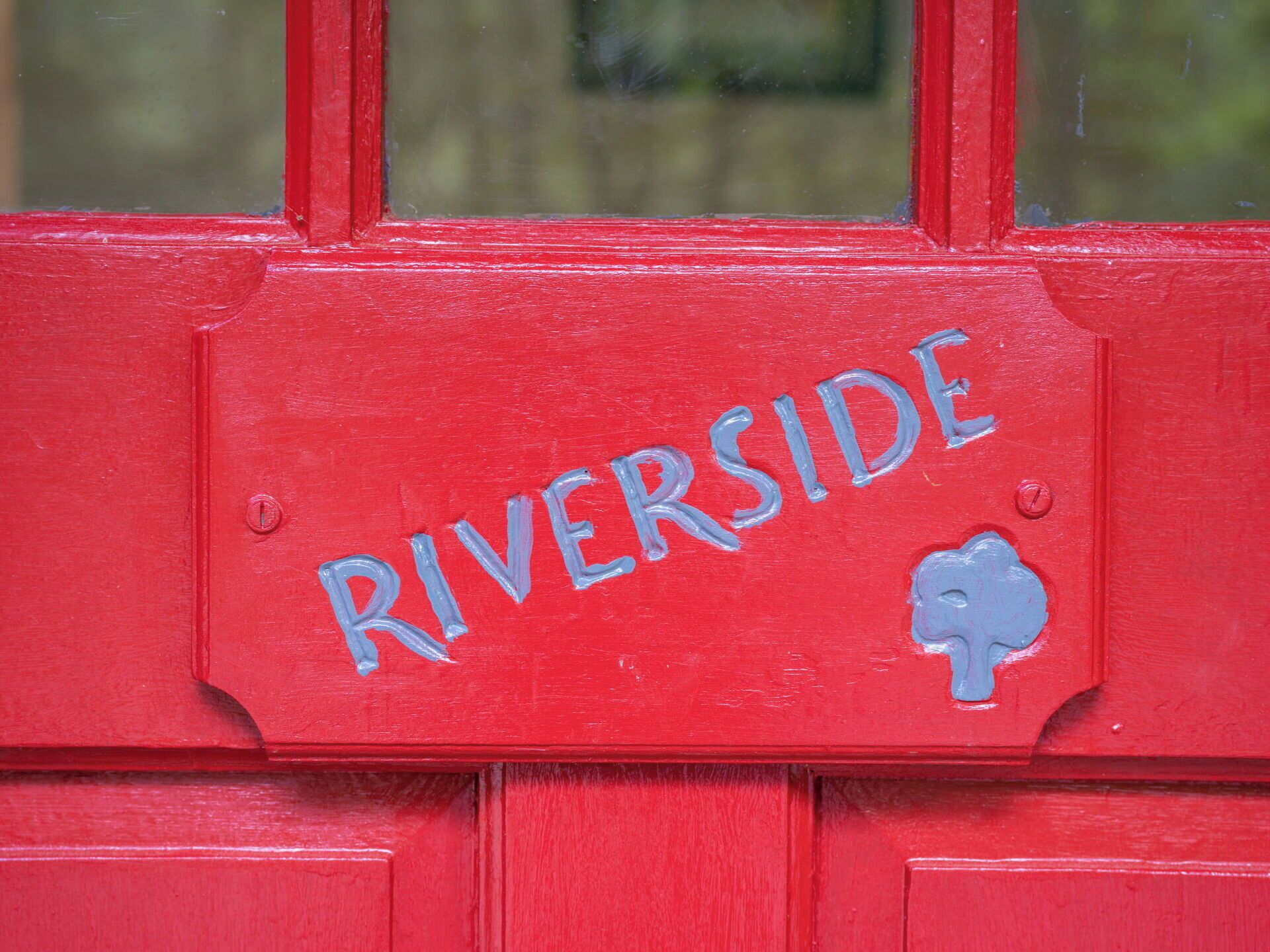 A red door with the word 'RIVERSIDE' painted in blue, a small cloud-like symbol, and two visible screws.