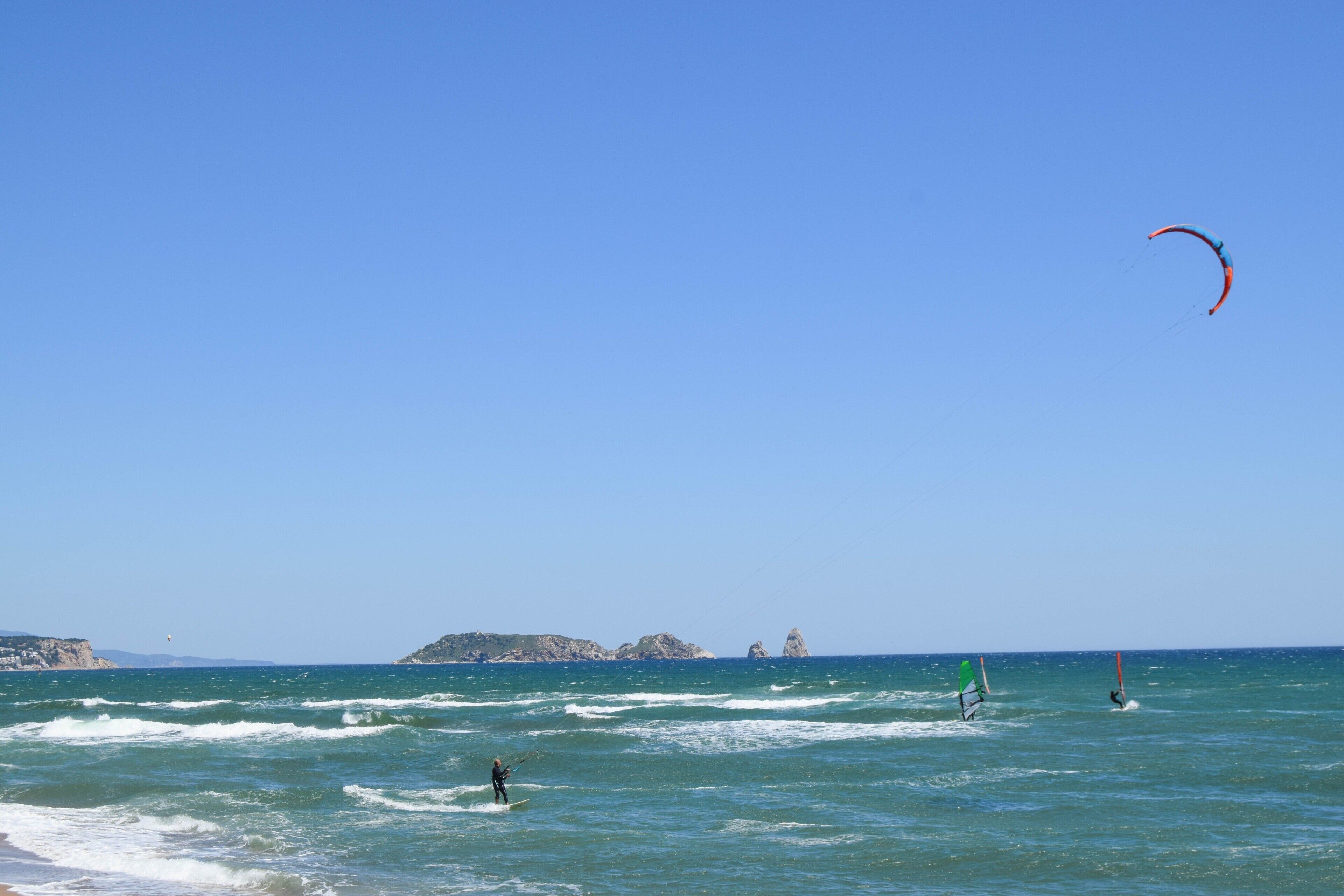 Uma praia com céu claro, ondas e uma pessoa praticando windsurf.