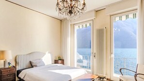 Ein Schlafzimmer mit Bett, einem Nachttisch aus Holz, einem Sessel, einer Chandelieber und Meerblick vom Balkon.