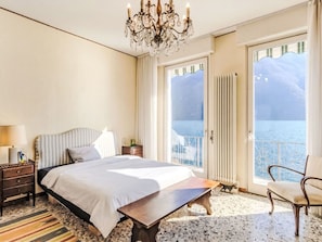 Een slaapkamer met een bed, een houten nachtkastje, een stoel, een kroonluchter en uitzicht op zee vanuit een balkon.