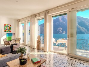 Een ruime kamer met een grote schuifglazen deur die leidt naar een balkon met uitzicht op een meer en bergen. Er staat een bank, een tafel met een boek en een bloempot.