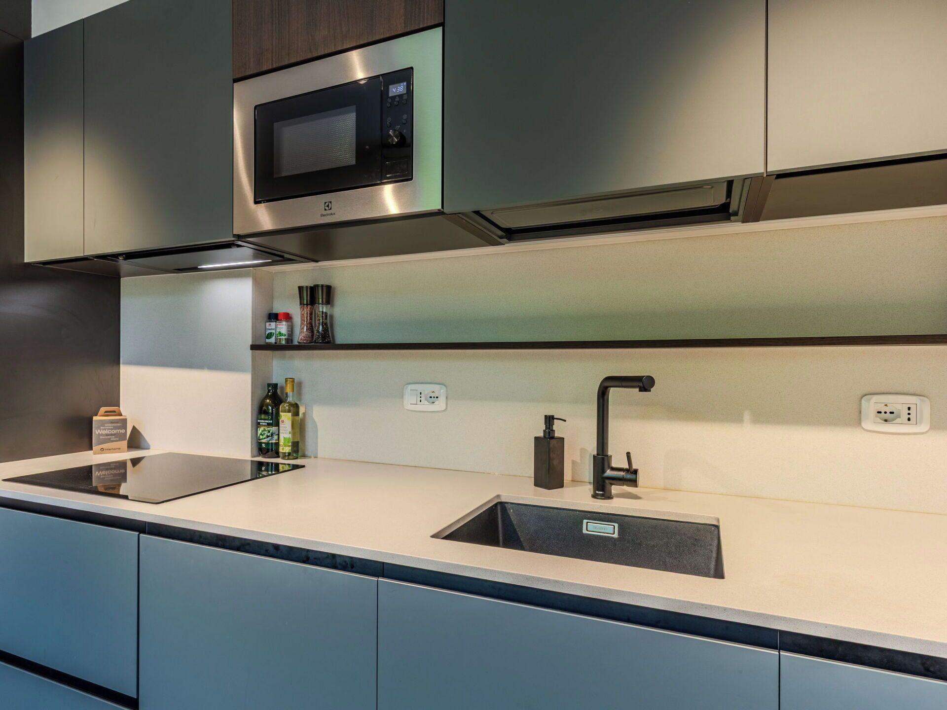 Una cocina moderna con microondas integrado, armarios de acero inoxidable, un lavamanos y una encimera con una botella de aceite de oliva.