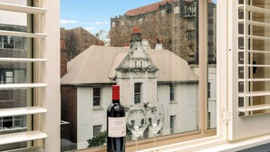 A window with a bottle of wine and two glasses on the sill, overlooking a building with a white facade and a red roof. - Hotel Emerald (Potts Point)