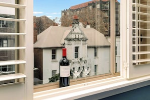 Une fenêtre sur le rebord de laquelle se trouvent une bouteille de vin et deux verres, donnant sur un immeuble à la façade blanche et au toit rouge.
