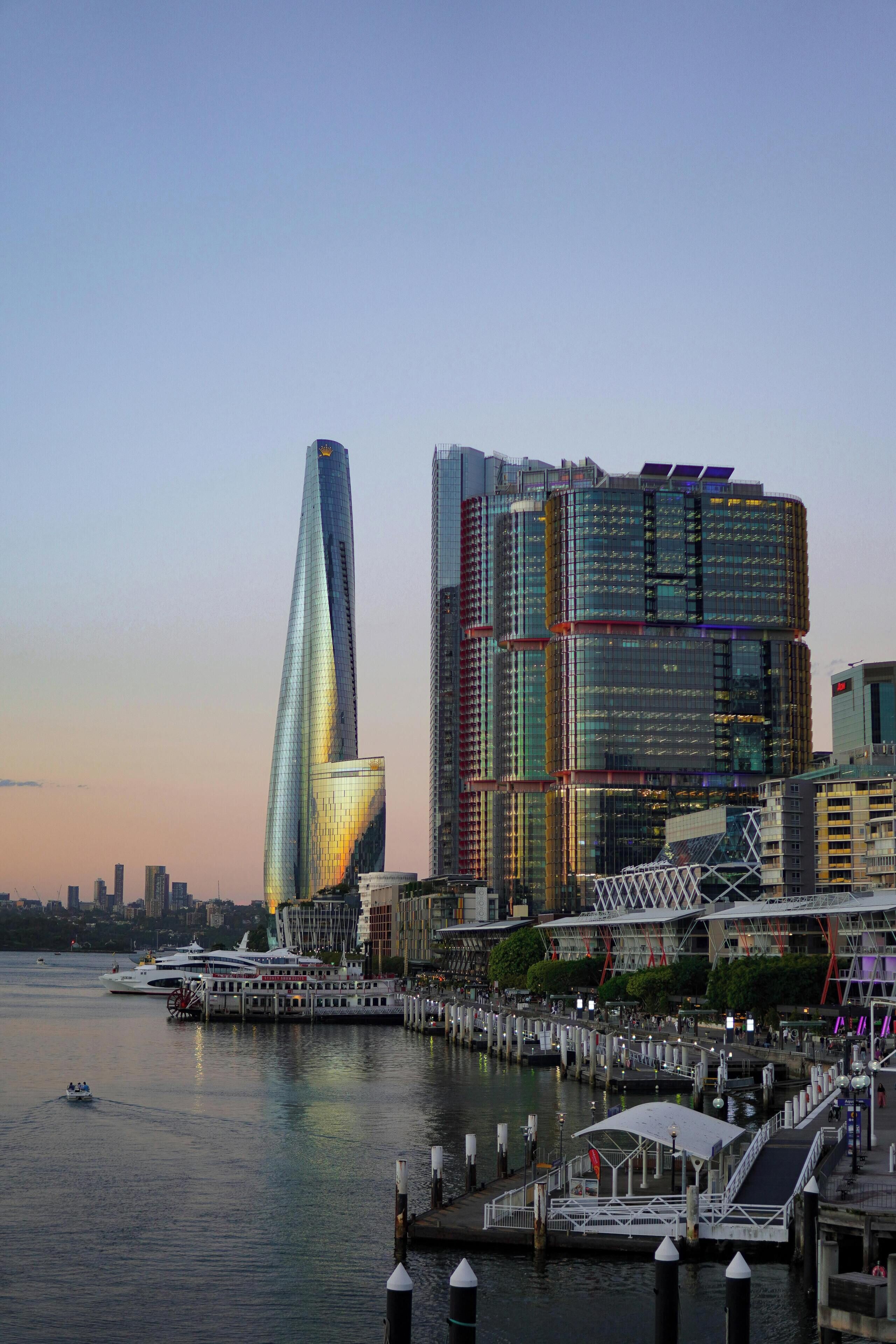 A waterfront cityscape with modern skyscrapers, a marina with boats, and a pedestrian bridge.