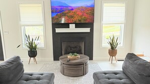 A living room with a flat-screen TV, a fireplace, two grey sofas, a round coffee table, and a ceiling fan. - East TN home with lake views & 2 kitchens (Morristown)