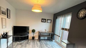 A living room with a flat-screen TV, a small table with a lamp, a potted plant, and a clock on the wall. - Stylish Stay in Emersons Green + WiFi + Garden (Emersons Green)