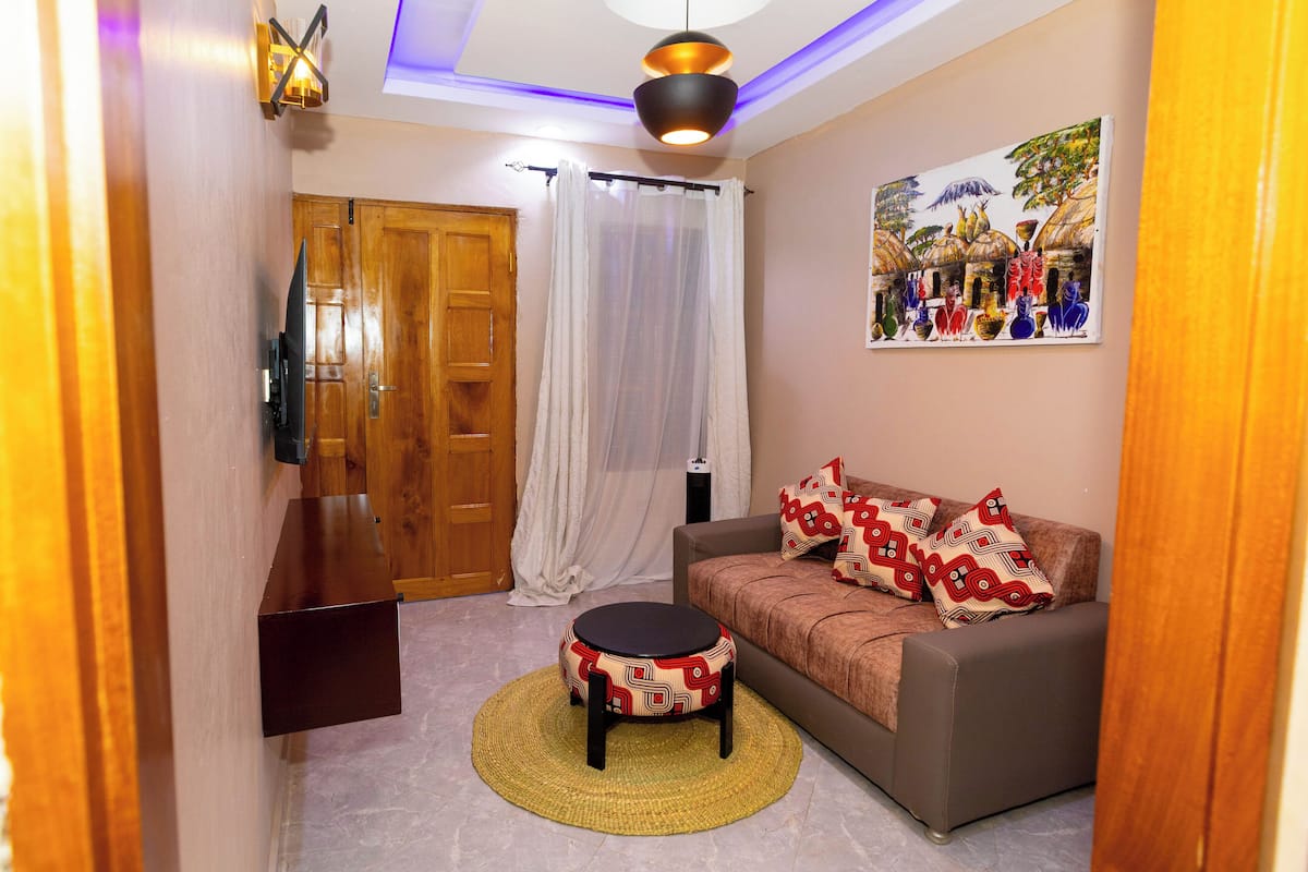 Una sala de estar con un sofá, una mesita redonda, un televisor montado en la pared, una puerta de madera y un cuadro colgado.