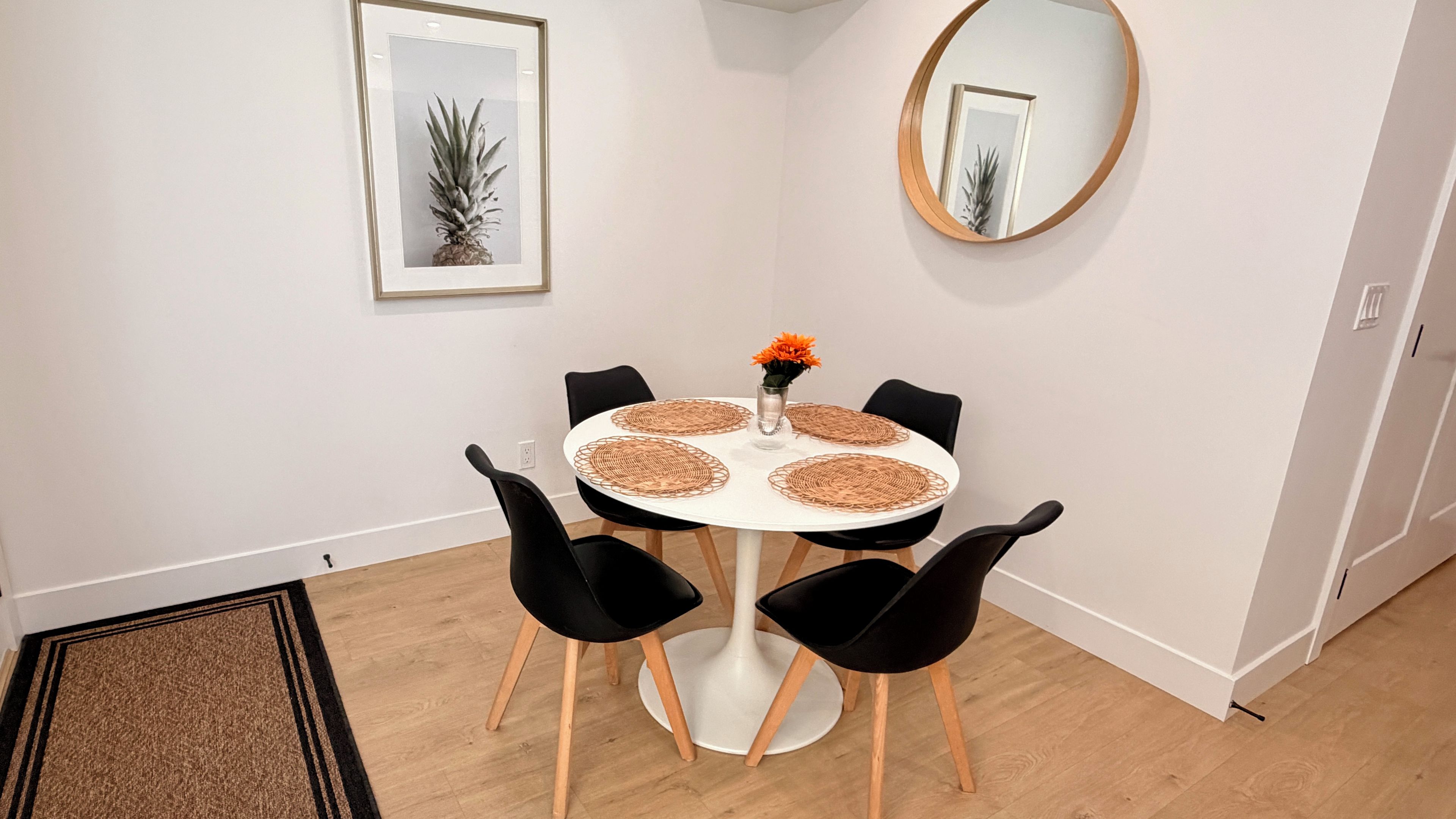 A dining area with a round table, black chairs, a vase with flowers, a framed picture on the wall, and a round mirror.