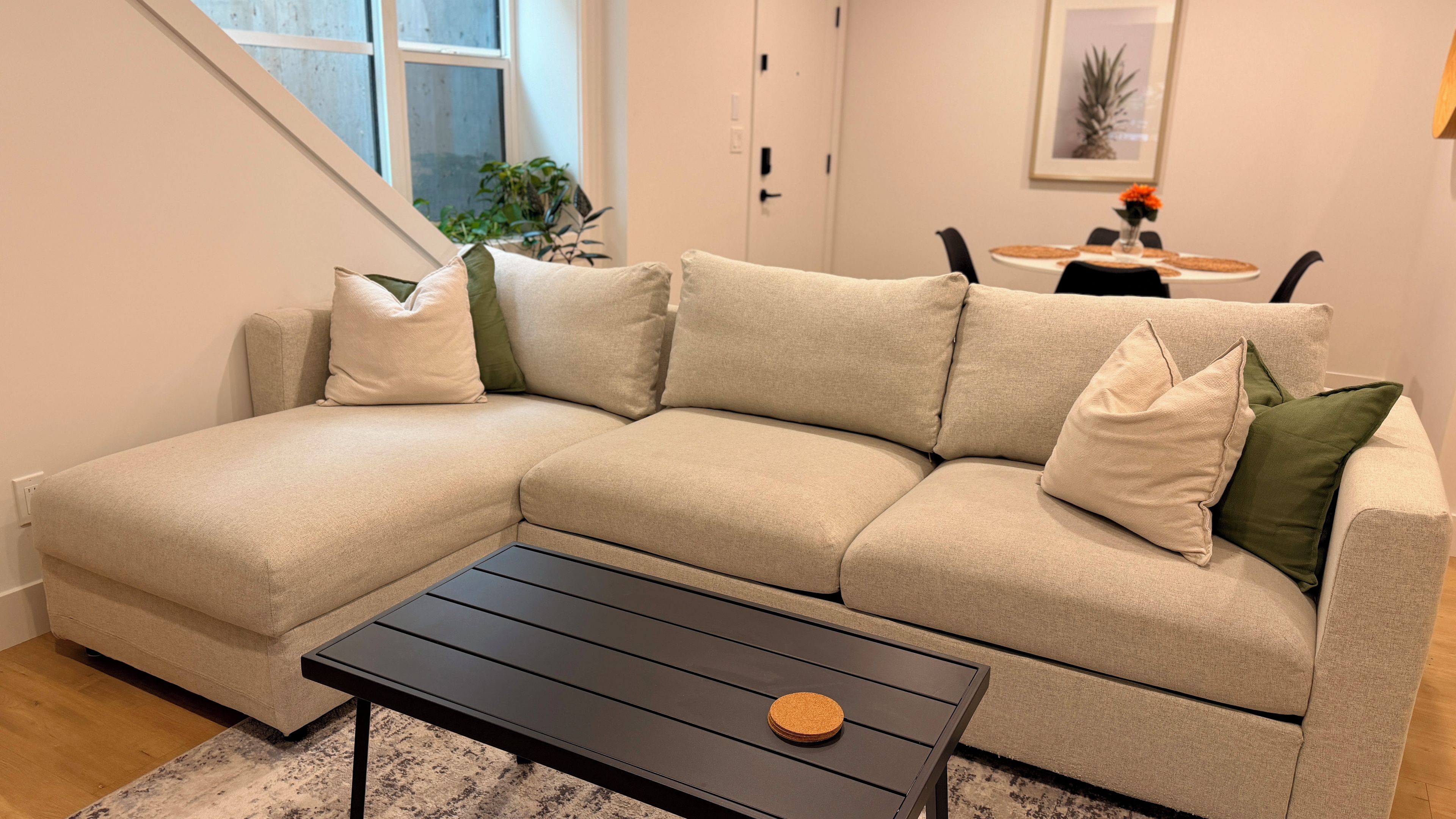 A beige sectional sofa with multiple cushions, a black coffee table with a cork coaster, and a dining area in the background.