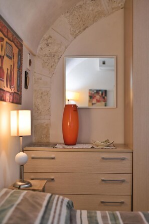 Una camera da letto con un letto, una lampada da comodino, una cassettiera e uno specchio. Sulla cassettiera, c'è un vaso.