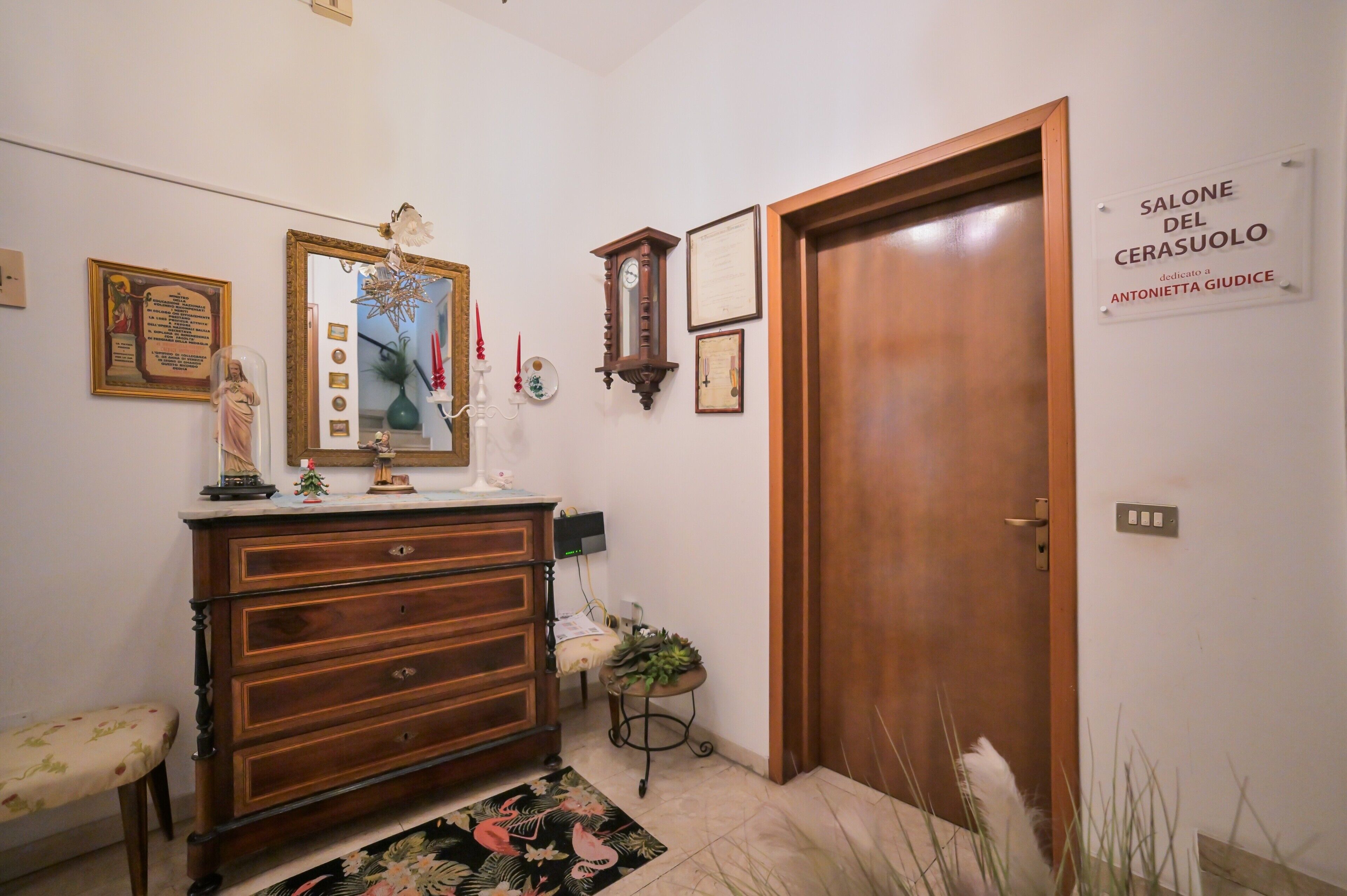 Quarto com penteadeira de madeira, espelho, relógio e porta com a inscrição 'SALONE DEL CERASUOLO'.