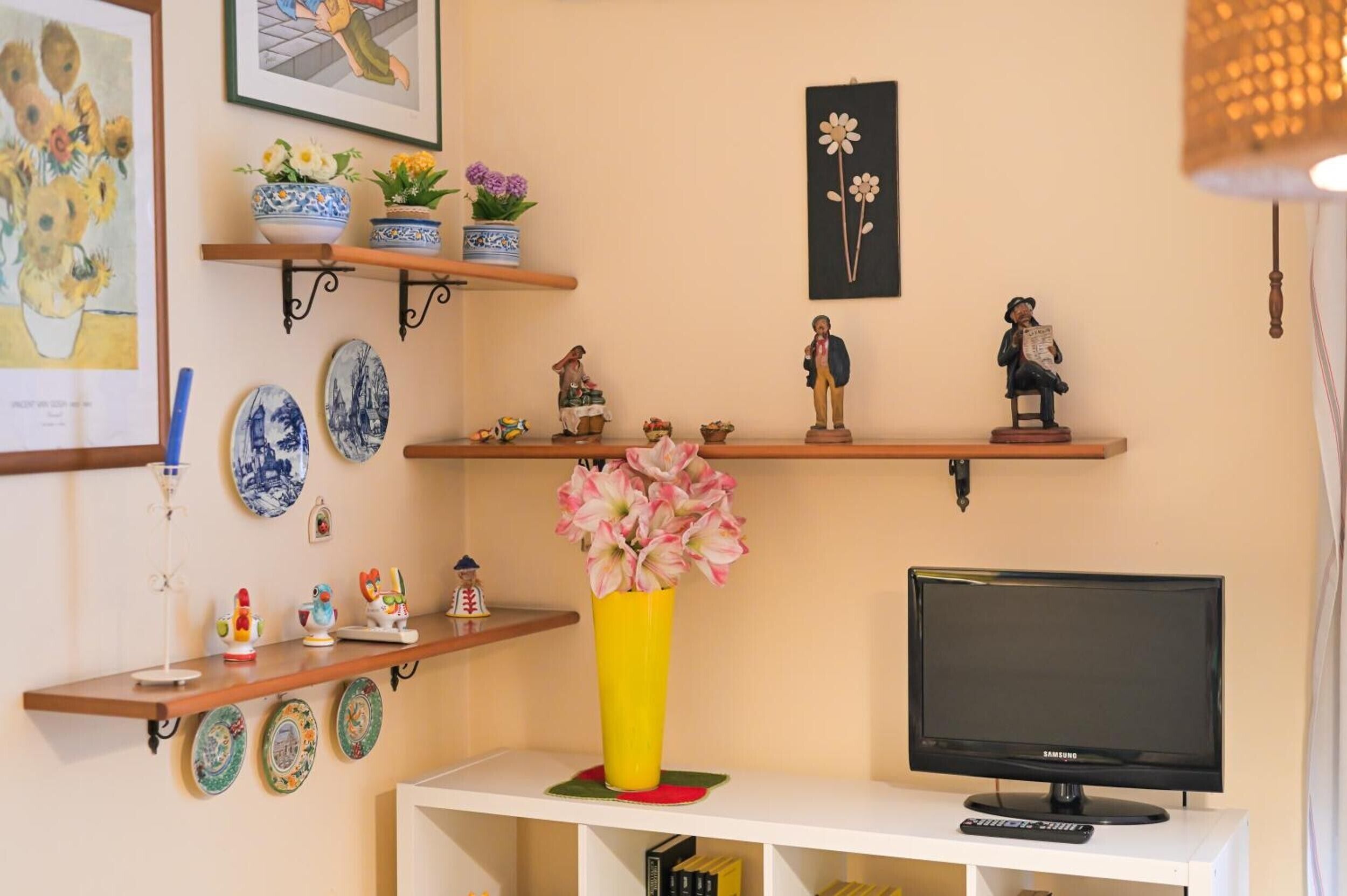 En stue med et fjernsyn, en gul vase med blomster og diverse dekorative genstande på hylder.