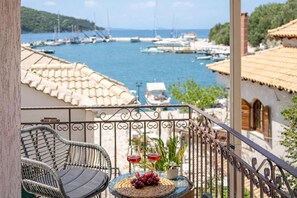 Un balcón con sillas de mimbre, una mesa pequeña con una copa de vino tinto y vista a un puerto deportivo con barcos.